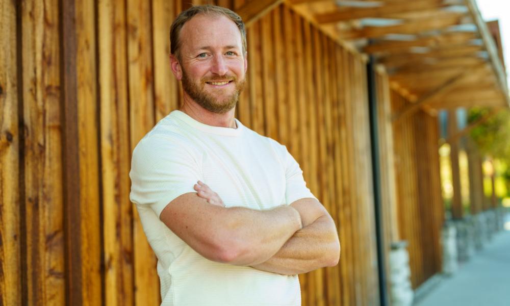 Travis in a white shirt with arms crossed standing in front of a wooden building